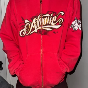 Röd huvtröja med tryck - Säljer en cool röd huvtröja med tryck på bröstet och en liten design på ärmen. Tröjan har dragkedja och är långärmad, perfekt för en avslappnad stil. Huvan ger extra komfort och stil. Perfekt för dig som vill sticka ut!