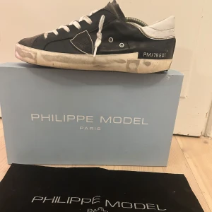 Svarta sneakers från Philippe Model - Snygga svarta sneakers från Philippe Model med vita snören och detaljer. Skorna har en unik design med en kombination av skinn och textilmaterial. Perfekta för en stilren look. Däremot finns det mindre skador på skorna, dm för fler bilder. Endast boxen medföljer!