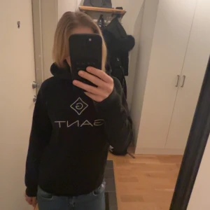 Svart hoodie från Gant - Säljer en svart hoodie från Gant med deras ikoniska logga på bröstet. Tröjan har en klassisk design med huva och en stor ficka framtill. Perfekt för en avslappnad stil. Har tyvärr ett litet håll på ena armen som man kan se på tredje bilden. Kan diskutera pris 