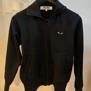 Svart jacka från Play - Snygg svart jacka från commes des garcons play med dragkedja och ett unikt hjärtmönster med ögon på bröstet och ryggen. Jackan har långa ärmar och en stilren design som passar perfekt för en cool look. Den är använd 1 gång. Nypris är: 2000