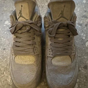 Grå Nike Air Jordans 4 i mocka - Snygga grå Nike Air Jordans 4 Kaws i mocka med klassisk snörning och ikonisk logga. Skorna har en stilren design med detaljer som ger dem en unik look. Perfekta för dig som vill ha något speciellt i garderoben. 