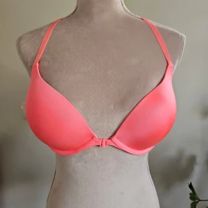 Korallfärgad bh från Hunkemöller - Säljer en snygg korallfärgad bh från Hunkemöller med unika banddetaljer på ryggen. Den har justerbara axelband och en slät design framtill. Perfekt för att ge en färgklick till din garderob!