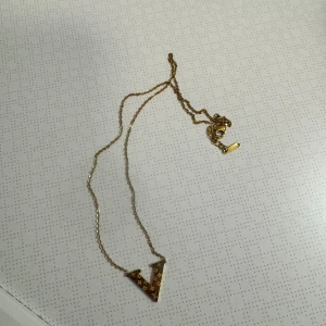 Guldhalsband med V-formad berlock med lv tryck på. -  guldhalsband med en V bokstav dekorerad med LV mönster. Halsbandet är 100% stainless steel det går och dusha med och använda dagligen.