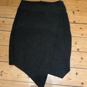 Grå kjol från Gina Tricot - Snygg grå kjol från Gina Tricot i en asymmetrisk design. Kjolen har en dragkedja baktill och är tillverkad i en mjuk blandning av polyester, viskos och elastan. Perfekt för en stilren look.             Längd 48 cm  Midja 66 cm   Dragkedjan i ryggen är något missfärgad/tappat färg