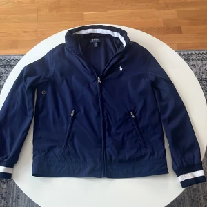 Mörkblå vindjacka från Ralph Lauren - Säljer en snygg mörkblå vindjacka från Ralph Lauren med luva i kragen. Liten i storleken,står 170 men jag skulle säga max 160cm