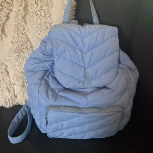 Blå quiltad ryggsäck - Säljer en snygg blå quiltad ryggsäck med dragkedja och snörning. Perfekt för att bära dina saker med stil. Ryggsäcken har justerbara axelremmar och en praktisk ficka framtill.