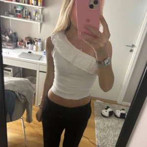 Vit oneshoulder topp med spetsdetalj - Säljer en stilren vit oneshoulder topp med vacker spetsdetalj längs axeln. Perfekt för en somrig look eller en utekväll. Toppen är tight och framhäver figuren på ett snyggt sätt.