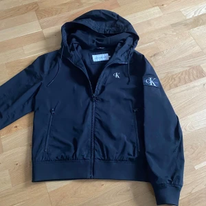 Svart vindjacka från Calvin Klein - Snygg svart vindjacka från Calvin Klein med huva och dragkedja. Jackan har långa ärmar och elastiska muddar. Perfekt för blåsiga dagar. Storlek M