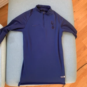 Tottenham Tröja - Säljer Tottenham tröja från Nike med dragkedja och klubbmärke på bröstet. Perfekt för fotbollsträning eller vardagsbruk. Tröjan har en stilren design med en liten etikett som visar 'Authentic Football'.