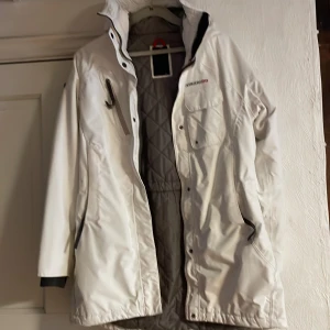 Vit parkajacka från Didriksons - Säljer en stilren vit parkajacka från Didriksons med quiltat foder och praktiska fickor. Jackan har en hög krage och justerbar huva, perfekt för kyliga dagar. Den har både dragkedja och knappar framtill för extra skydd mot väder och vind.