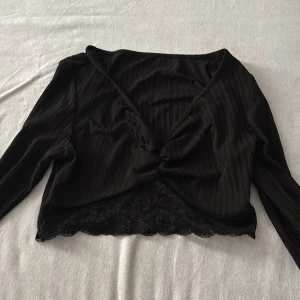 Svart långärmad topp med spetsdetaljer. - Snygg svart långärmad topp med spetsdetaljer i nederkant. Toppen har en omlott-design och är ribbad för en stilren look. Perfekt för en chic och elegant stil.