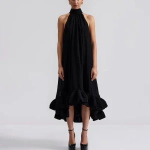 Svart ärmlös klänning med volangdetaljer från By Malina - Sabrina Maxi Elegant svart klänning med hög hals och volangdetaljer vid fållen. Den är ärmlös och har en lös passform som ger ett stilrent och sofistikerat intryck. Perfekt för speciella tillfällen.