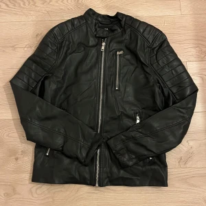 Svart skinnjacka  - Snygg svart skinnjacka med coola detaljer som dragkedjor och quiltade axlar. Perfekt för en stilren look. Jackan har en klassisk passform och långa ärmar. Mycket bra skick. 