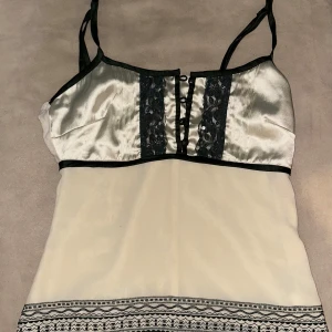 Vit/beige och svart linne från Vero Moda - Snyggt linne från Vero Moda i vitt/beige och svart med spetsdetaljer framtill. Linnet har smala axelband och en dekorativ kant längst ner. Perfekt för en stilren look. Dragkedja på sidan för en mer definierad look