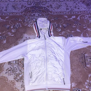 Vit jacka från Moncler - Säljer en snygg vit jacka från Moncler med dragkedja och huva. Jackan har ribbade detaljer och Moncler-logga på ärmen. Perfekt för en stilren look.