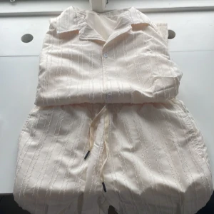 Beige skjorta och shorts set - Snyggt set med beige skjorta och matchande shorts. Skjortan har knappar och ett subtilt randigt mönster. Shortsen har en bekväm dragsko i midjan. Perfekt för en avslappnad stil.