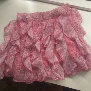 Rosa blommig volangkjol - Charmig rosa kjol med blommigt mönster och volanger. Perfekt för en somrig look med sin korta längd och lekfulla design.