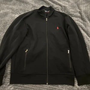 Svart zip tröja från Polo Ralph Lauren - Säljer en stilren svart sweatshirt från Polo Ralph Lauren med dragkedja och röd broderad logotyp på bröstet. Skick 10/10