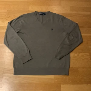 Beige/grå tröja från Polo Ralph Lauren - Säljer en stilren beige/grå tröja från Polo Ralph Lauren med klassisk v-ringning och broderad logotyp på bröstet. Tröjan har långa ärmar och ribbade muddar vid ärmslut och nederkant. Perfekt för en avslappnad och elegant look. 9,5 av 10 i skick