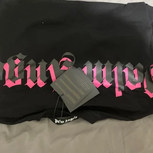 Svart t-shirt från Palm Angels - Säljer en svart t-shirt från Palm Angels med rosa gotisk text på bröstet. T-shirten har en klassisk rund halsringning och korta ärmar. Perfekt för en stilren och edgy look.