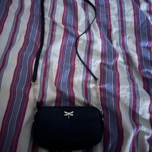 Svart axelväska med dragkedja - Snygg svart axelväska med en elegant dragkedja och en dekorativ detalj i form av en liten slända på framsidan. Väskan har en lång axelrem i kombination av kedja och material, perfekt för att bära över axeln.