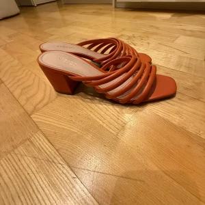 Orange klackskor från Arezzo - Snygga orange klackskor från Arezzo med flera remmar över foten. Tillverkade i Brasilien, dessa skor har en fyrkantig tå och en stabil klack som ger en modern touch. Perfekta för att lägga till en färgklick i din outfit. kom med prisförslag 
