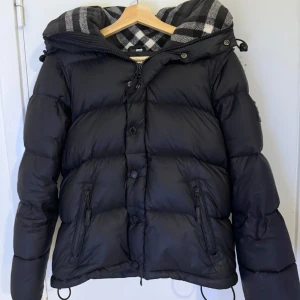 Burrbery Jacka  - Säljer en snygg svart dunjacka från Burberry med klassiskt rutigt foder i huvan. Jackan har dragkedja och knappar framtill samt två praktiska fickor med dragkedja. Perfekt för kyliga dagar!