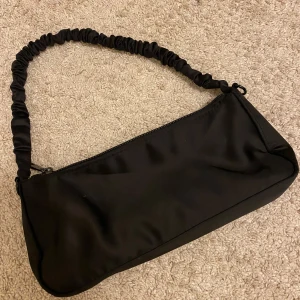 Svart axelväska med rynkad rem - Snygg svart axelväska med en unik rynkad rem och dragkedja upptill. Perfekt för att bära dina nödvändigheter med stil. Enkel och elegant design som passar till många tillfällen.