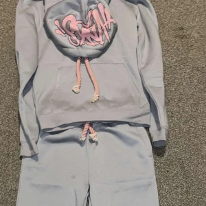 Ljusblå hoodie med rosa detaljer - Säljer en ljusblå hoodie med rosa snören och tryck på huvan. Tröjan har en stor ficka framtill och är perfekt för en avslappnad stil. Matchande byxor med elastisk midja och rosa snöre ingår. Den är helt oanvänd och i storlek M