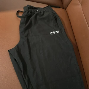 Svarta byxor från Shein - Säljer ett par svarta byxor från Shein med elastisk midja och snörning. Byxorna har en avslappnad passform och är perfekta för en bekväm stil. Märket 'AUSSUP' är tryckt på framsidan.