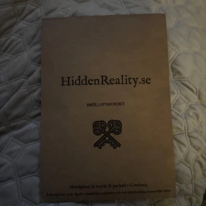 Bröllopsmordet från HiddenReality.se - Ett spännande mordgåtespel från HiddenReality.se med titeln 'Bröllopsmordet'. Perfekt för en mystisk spelkväll där du och dina vänner kan lösa gåtor tillsammans. Förpackningen är stilrent designad i brunt med två korsade nycklar som symbol.