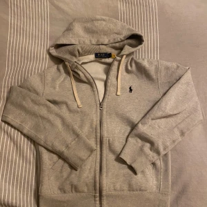 Grå hoodie från Polo Ralph Lauren - Säljer en stilren grå hoodie från Polo Ralph Lauren med dragkedja och broderad logga på bröstet. Tillverkad i mjuk bomullsblandning med huva och snören. Perfekt för en avslappnad look.
