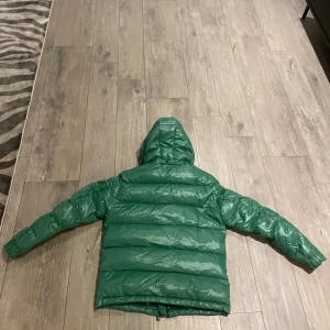 Grön dunjacka från Moncler Maya  - Säljer en snygg grön dunjacka från Moncler med huva och dragkedja. Jackan har långa ärmar och en broderad logotyp på ärmen. Perfekt för kyliga dagar. Bild på årder bekräftelse finns vid intresse 