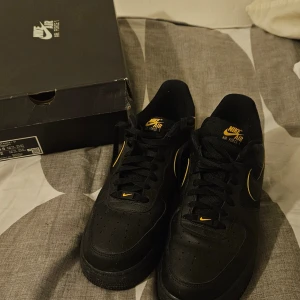 Nike Air Force 1 '07 i svart och guld - Säljer ett par Nike Air Force 1 '07 i svart med detaljer i guld. Skorna har klassisk snörning och är tillverkade i skinn. Perfekta för en stilren look.