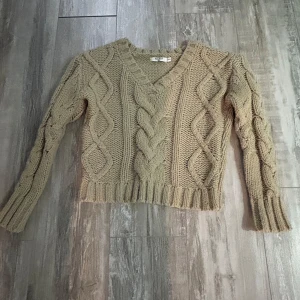 Beige stickad tröja från NA-KD - Säljer en mysig beige stickad tröja från NA-KD med flätmönster och v-ringning. Tröjan har långa ärmar och ribbade kanter, perfekt för kyliga dagar. Använd få gånger och är i mycket bra skick!