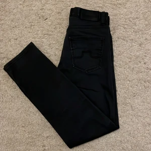 Svarta byxor från J.Lindeberg - Snygga svarta byxor från J.Lindeberg med en stilren design. Byxorna har en diskret logga och är perfekta för en modern look. De har en klassisk passform och är tillverkade i ett bekvämt material.