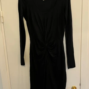 Svart klänning från Designers Remix Collection - Elegant svart klänning från Designers Remix Collection i storlek XS. Klänningen har långa ärmar och en stilren design med en knutdetalj framtill som ger en smickrande silhuett. Perfekt för en chic look.