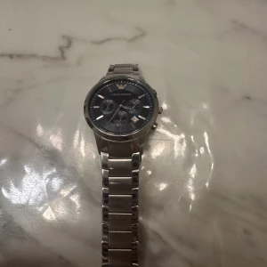 Silverfärgad klocka från Emporio Armani - Snygg och stilren klocka från Emporio Armani i silverfärg. Den har en metallarmband och en mörk urtavla med flera små urtavlor för extra funktionalitet. Perfekt för den som vill ha en elegant accessoar.