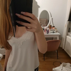 Vit linne med spetsdetaljer från Amisu - Säljer ett stilrent vitt linne från Amisu med vackra spetsdetaljer längs halsringningen. Linnet har smala skjusterbara axelband och är perfekt för en elegant look. Passar bra till både jeans och kjol.