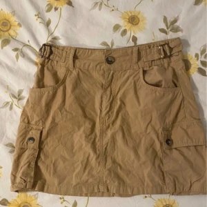 Beige kjol från Bershka - Säljer en beige kjol från Bershka i medium waist-modell. Kjolen har praktiska fickor med knappar på sidorna och en bekväm passform. Perfekt för en avslappnad stil.