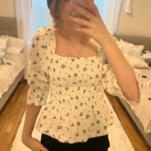 Blommig blus med puffärmar - Charmig blommig blus med puffärmar och smockdetaljer. Den har en fyrkantig urringning och är perfekt för en romantisk look. Blusen är vit med små blå blommor och har en lätt volang vid nederkanten.