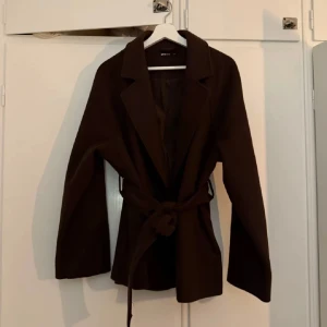 Brun kappa med knytband - Snygg brun kappa med knytband i midjan. Perfekt för höst och vår. Den har långa ärmar och en klassisk krage. En stilren och tidlös design som passar till många tillfällen.