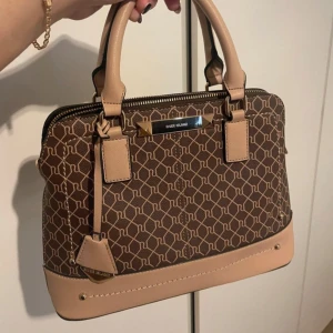Brun och beige handväska från River Island - Snygg handväska från River Island i brunt och beige med ett mönster. Väskan har en dragkedja upptill och bekväma handtag. Perfekt för både vardag och fest! Använd endast en gång! Köpt för 58 pund motsvarar 800kr. Axelrem medföljs. Pris kan diskuteras!