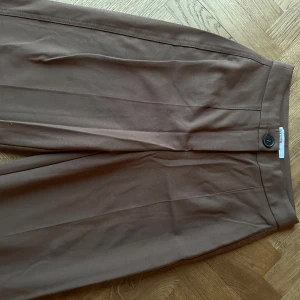 Bruna kostymbyxor - Snygga bruna kostymbyxor med hög midja och raka ben. Byxorna har en knappdetalj både fram och bak, vilket ger en stilren look. Perfekta för en chic och elegant stil.