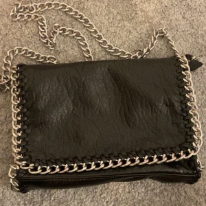 Svart axelväska med kedjedetaljer från Tiamo - Snygg svart axelväska från Tiamo med silverfärgade kedjedetaljer runt kanterna och som axelrem. Den är i väldigt bra skick, endast använd vid få tillfällen!