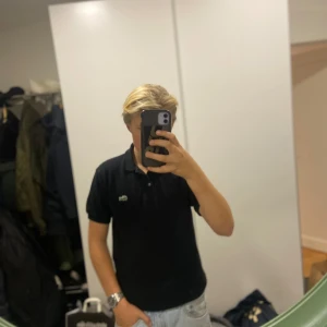 Svart pikétröja från Lacoste - Säljer en klassisk svart pikétröja från Lacoste med den ikoniska krokodilloggan på bröstet. Tröjan har korta ärmar och en stilren krage med knappar. Perfekt för en avslappnad men ändå stilren look. Nypris 1100! Mitt pris går att diskuteras! 