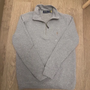 Ralph Lauren Quarter Zip - Säljer en stilren grå tröja från Polo Ralph Lauren i mycket bra skick. Den har en dragkedja vid halsen och är långärmad, perfekt för höst och vinter. Tröjan är i en mjuk bomullsmix och har en liten broderad logga på bröstet. Passar både till vardags och lite finare tillfällen. Skick 10/10 oanvänd