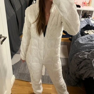 Vit fleece onesie med huva - Mysig vit onesie i fleece med huva och dragkedja framtill. Den har ett hjärtmönster som ger en söt touch. Perfekt för kalla dagar hemma.