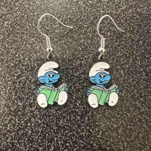 Örhängen med smurfmotiv - Charmiga örhängen med smurfmotiv i blått, grönt och vitt. Perfekta för att ge en lekfull touch till din stil. De är lätta och bekväma att bära hela dagen. Krokarna är inköpta som nickelsäkra.