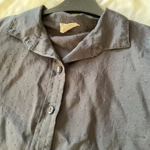 Svart skjorta från Miss Dee - Säljer en stilren svart skjorta från Mister Dee. Skjortan har långa ärmar och knappar framtill. Perfekt för både vardag och festliga tillfällen. Den är i bra skick och har en klassisk krage. Passar utmärkt till höst- och vårgarderoben.